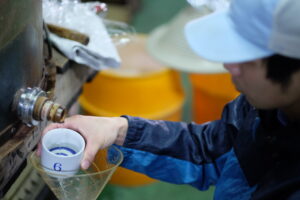 Brewery worker sampling sake during quality inspection at Mitsutake Brewery